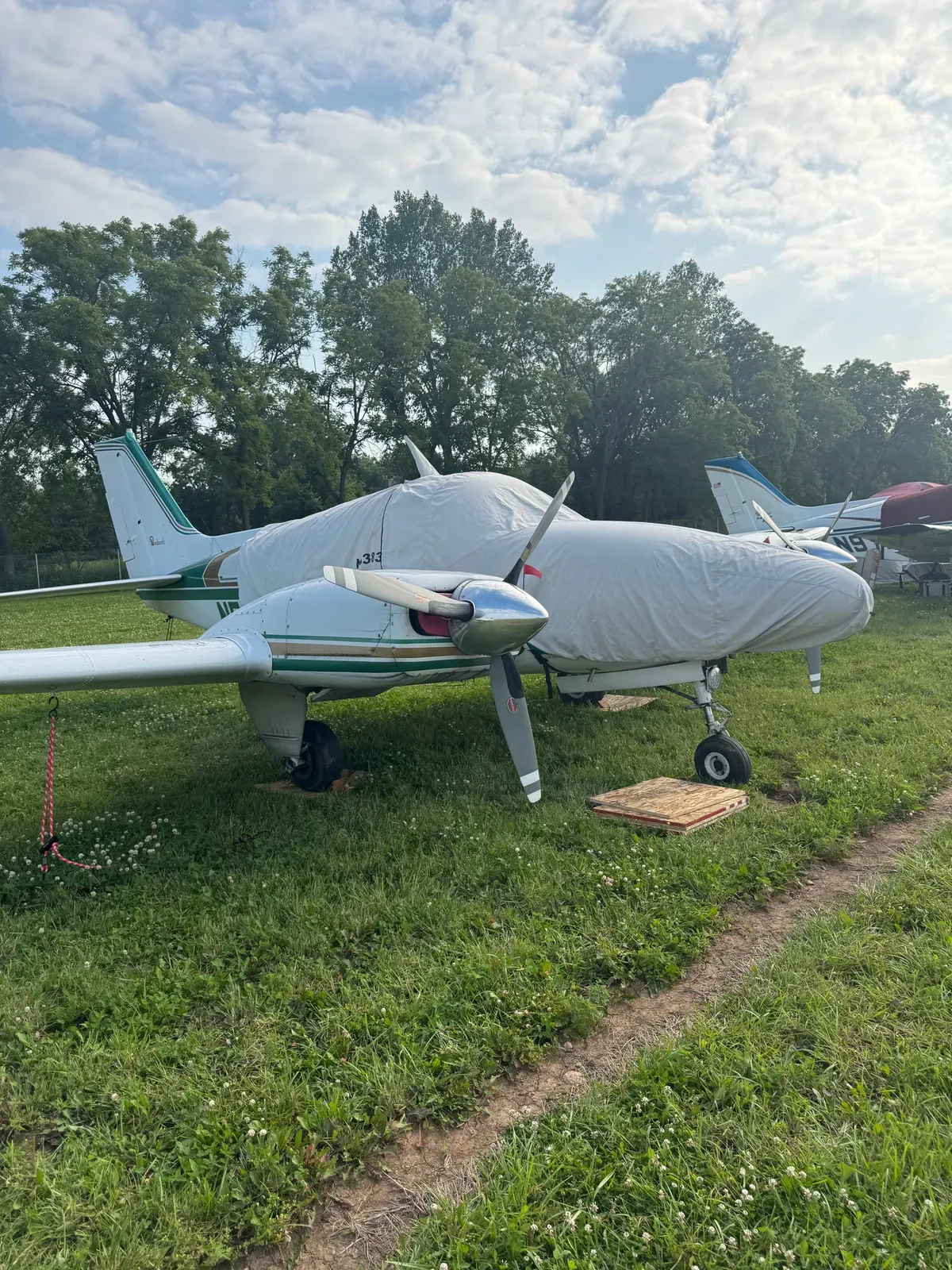 A Beechcraft Baron tied down on the grass at Oshkosh with cowl covers on and a wood board under the nosewheel